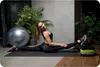 A woman is doing a split stretch on a yoga mat in a home gym setting. There is a large exercise ball and plants in the background. Voir les images du produit