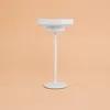 A white table lamp with a circular base and a disc-shaped top is shown against a light peach background. Produktbild-Vorschau