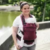 A woman is carrying a baby in a carrier in an outdoor park setting. Produktbild-Vorschau