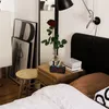 A bedside table with an alarm clock, a rose in a vase, and a patterned box next to a bed. Produktbild-Vorschau