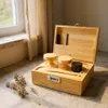 A wooden storage box with a combination lock, open to reveal two glass jars with wooden lids, a black container, and some accessories. The box is placed on a textured surface next to a window with sheer curtains and some dried herbs. Produktbild-Vorschau