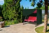 A red swing set is covered by a dark green protective cover in an outdoor garden setting. Náhľad obrázku produktu