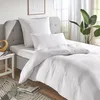 A neatly made bed with white bedding, pillows, and a duvet, set against a grey headboard and a window with sheer curtains and a plant. Produktbild-Vorschau