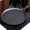 A cast iron frying pan is shown on a black reflective surface, with a wooden object and a plant in the background. Produktbild-Vorschau
