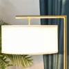 A close-up of a modern floor lamp with a white drum shade and a gold metal frame, set against a teal curtain and some greenery. Náhľad obrázku produktu