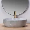 A beige oval basin sink with a gold faucet in front of a round mirror. Náhled obrázku produktu