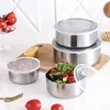 A set of stainless steel food containers with clear lids, arranged on a wooden cutting board and a white surface, with some food items and a lemon slice visible. Produktbild-Vorschau