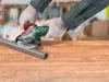 A person wearing gloves is using a cordless angle grinder to cut a metal bar on a wooden surface. Produktbild-Vorschau