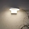 A modern, circular wall light fixture is mounted on a textured, light-colored wall. The light is illuminated, casting a soft glow and a shadow of a plant on the wall. Produktbild-Vorschau