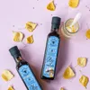 Two bottles of oil are arranged diagonally on a pink background, surrounded by fallen yellow leaves and scattered black seeds. A small glass jar with a spoon and some white substance is also present. Produktbild-Vorschau