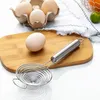 A stainless steel egg separator is placed on a wooden cutting board next to a single egg. In the background, an egg carton with three eggs is visible. Produktbild-Vorschau