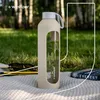 A glass water bottle with a tan silicone sleeve is placed on a picnic blanket outdoors, with grass and trees in the background. Sunglasses and a book are also visible. Voir les images du produit