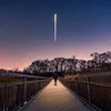 A person stands on a wooden bridge at night, looking up at a streak of light in the starry sky. Náhľad obrázku produktu