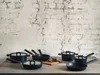 A collection of black cookware is arranged on a wooden table against a textured white brick wall. Podgląd zdjęcia produktu