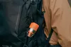 A close-up of an orange and clear portable air pump attached to a black backpack, worn by a person in a tan jacket. Podgląd zdjęcia produktu
