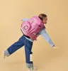 A young girl wearing a pink backpack and jeans is balancing on one leg against a plain background. Náhled obrázku produktu