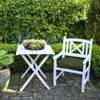 A white garden table and chair set with a cushion on the chair, placed on a paved patio surrounded by lush green bushes and trees. Náhled obrázku produktu