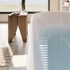 A modern bathroom scene with a freestanding bathtub filled with water, a wooden stool with a diffuser and soap, and a large window overlooking a beach. Náhled obrázku produktu