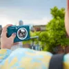 A person is holding a teal Canon Zoemini S2 camera, taking a selfie with a blurred background of trees and buildings. Anteprima dell’immagine prodotto