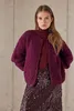 A woman wearing a burgundy turtleneck and a textured burgundy cardigan over a sequined burgundy skirt, standing against a concrete wall. Produktbild-Vorschau