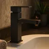 A black modern faucet is shown in a bathroom setting, positioned over a white sink. The background features a textured wall and a plant. Podgląd zdjęcia produktu