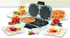 Unold Waffelautomat offen auf weißem Tisch, umgeben von Waffeln, Früchten, Sirup, Sahne und Beeren. Produktbild-Vorschau