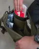 A person is placing a red and white item into a green backpack. Produktbild-Vorschau