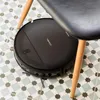 A black robot vacuum cleaner is cleaning the floor under a chair. Náhled obrázku produktu
