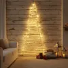 A rustic, wall-mounted Christmas tree made of wooden sticks and adorned with fairy lights, set against a wooden plank wall in a cozy living room setting with a sofa and gifts. Podgląd zdjęcia produktu