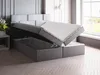 A modern bedroom with a grey upholstered bed frame, featuring a lift-up storage compartment and a mattress. The room has a large window with natural light and a wooden floor. Produktbild-Vorschau