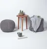A knitted pouf, a small wooden table with a cactus, a basket with a blanket and magazines, and a mug on a book are arranged on a white surface against a white background. Náhled obrázku produktu