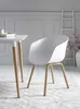 A white modern chair with wooden legs is placed next to a white table on a patterned rug. Náhľad obrázku produktu