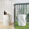 A split image showing a white ceramic stool in two different settings: indoors on a wooden floor and outdoors on grass, both with a railing in the background. Anteprima dell’immagine prodotto
