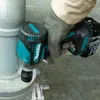 A person wearing a work glove is using a Makita cordless impact wrench to tighten a bolt on a metal pipe fitting. Produktbild-Vorschau