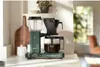 A coffee maker on a kitchen counter with a cutting board and a plant in the background. Náhled obrázku produktu