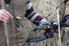 A person is holding an arrow and a bow in an outdoor setting with dry grass and trees. Anteprima dell’immagine prodotto