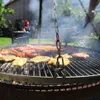A large round grill with various meats and seafood cooking on it, set in an outdoor backyard setting with people in the background. Podgląd zdjęcia produktu