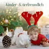 A young boy wearing reindeer antlers and a red sweater smiles as a small white dog nuzzles him. A Christmas tree with lights and ornaments is in the background, along with gift-wrapped boxes. A pinecone-shaped candle with a lit flame is in the foreground. Produktbild-Vorschau