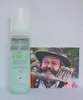 A bottle of Goldwell Dualsenses Curls & Waves Hydrating Serum Spray is placed next to a postcard featuring a man with a large, decorated beard and hat. Náhled obrázku produktu