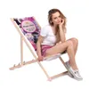 A woman is sitting on a foldable wooden deck chair with a floral pattern and text on it. Produktbild-Vorschau