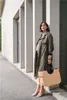 A woman stands leaning against a building, holding a large shopper bag. Produktbild-Vorschau