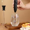 A hand holding a black electric milk frother over a glass of water, with a stand for the frother and a cupcake in the foreground. Produktbild-Vorschau