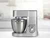 A silver kitchen robot is displayed on a white surface with a blurred background. Produktbild-Vorschau