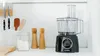 A black Bosch food processor sits on a white countertop next to a jar of flour and two wooden salt and pepper grinders. Kitchen knives are mounted on the wall behind. Produktbild-Vorschau