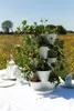 A tiered strawberry planter sits on a table covered with a white tablecloth, set in a field of yellow flowers under a clear blue sky. A teapot and bottles are also on the table. Produktbild-Vorschau