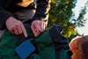 A person is shown reaching into a green and black backpack, with a small blue case visible. A brown dog is partially visible in the background. Produktbild-Vorschau