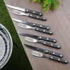 A set of steak knives and forks laid out on a wooden table next to a grill. Voir les images du produit