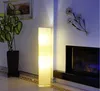 A tall, rectangular floor lamp with a beige paper shade stands in a room next to a large potted plant and a fireplace. Produktbild-Vorschau