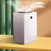 A white air purifier is placed on a wooden shelf, with green curtains in the background and a light orange wall behind it. A faint mist is visible above the air purifier. Produktbild-Vorschau