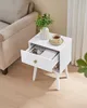 A white nightstand with an open drawer and a cup of tea on top, next to a couch. Náhled obrázku produktu